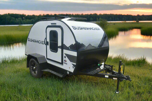 A Braxton Creek Bushwhacker travel trailer RV with a black and white exterior, parked in a natural setting with grass and water in the background.