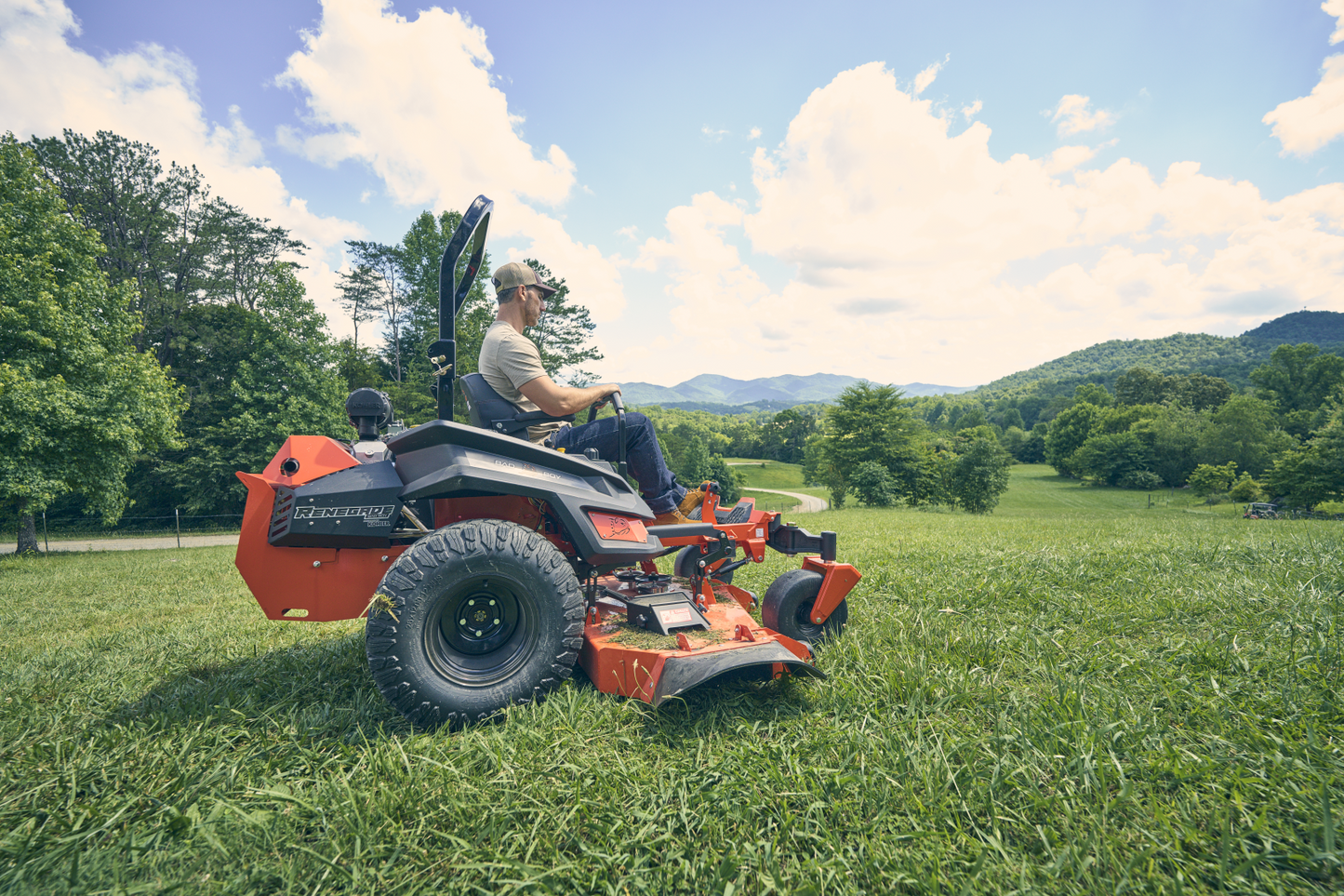 Man operating a lawn mower in a grassy field with trees and mountains in the background