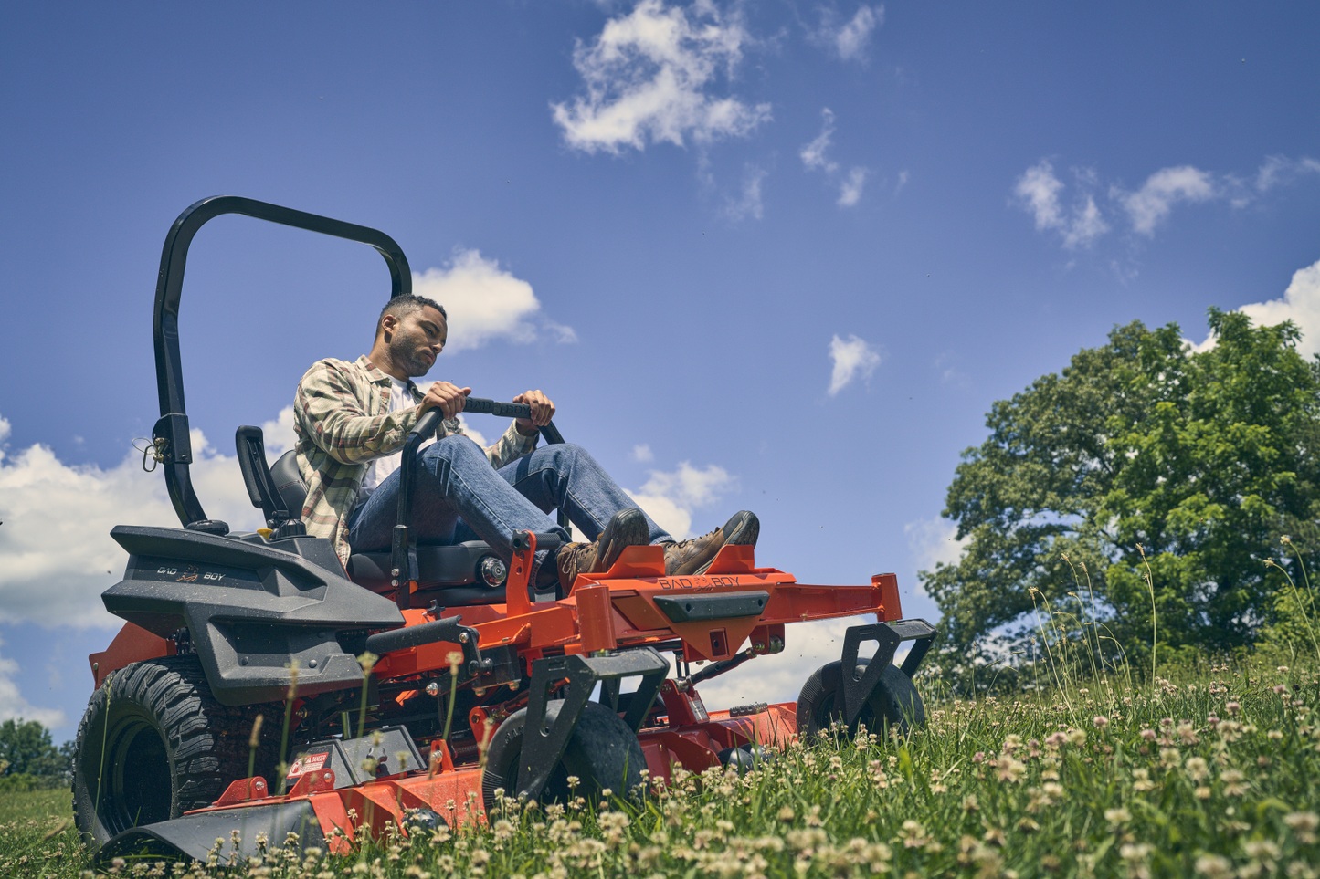 Man operating a red lawn mower in a grassy field with blue sky and trees in the background