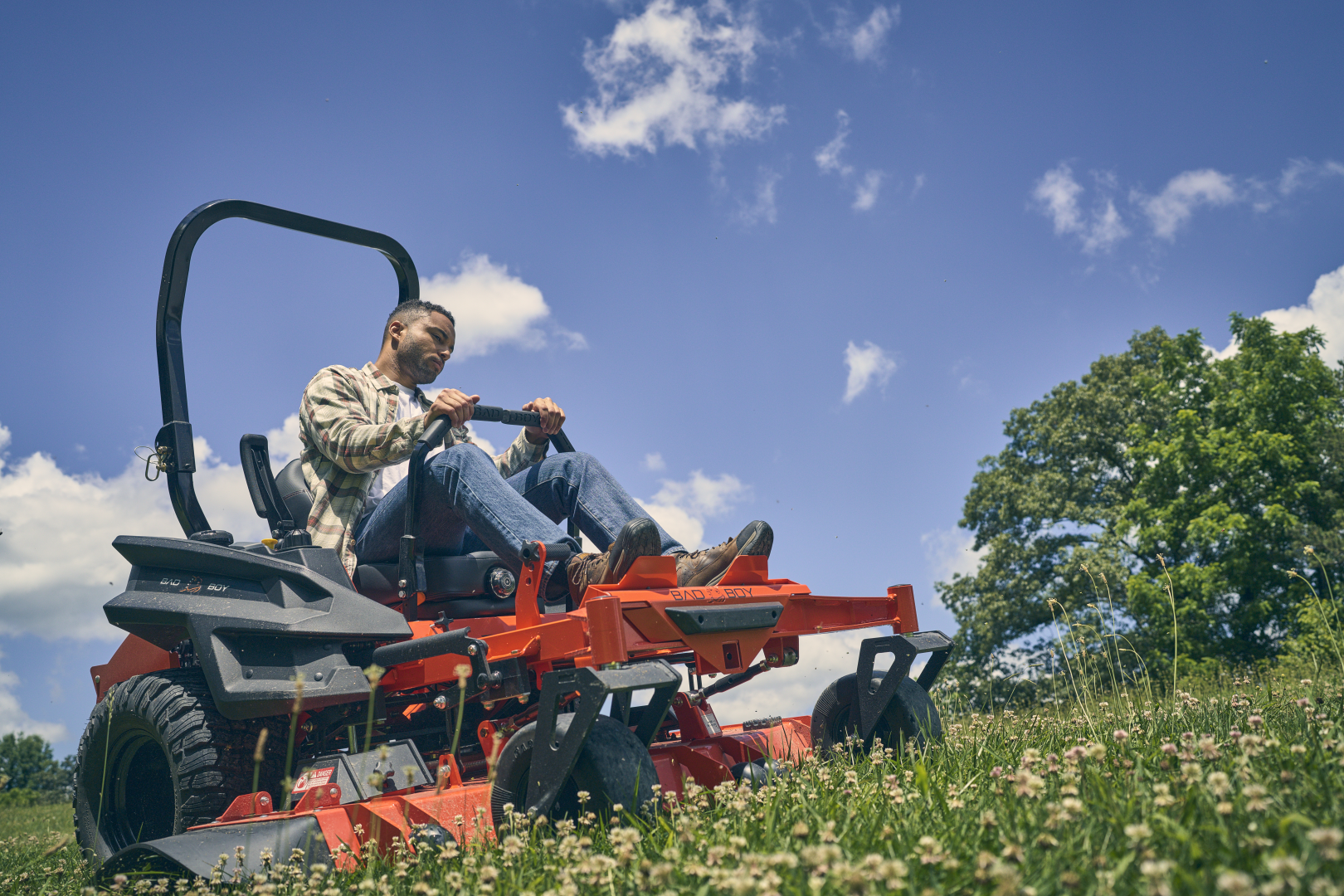 Man operating a red lawn mower in a grassy field with blue sky and trees in the background