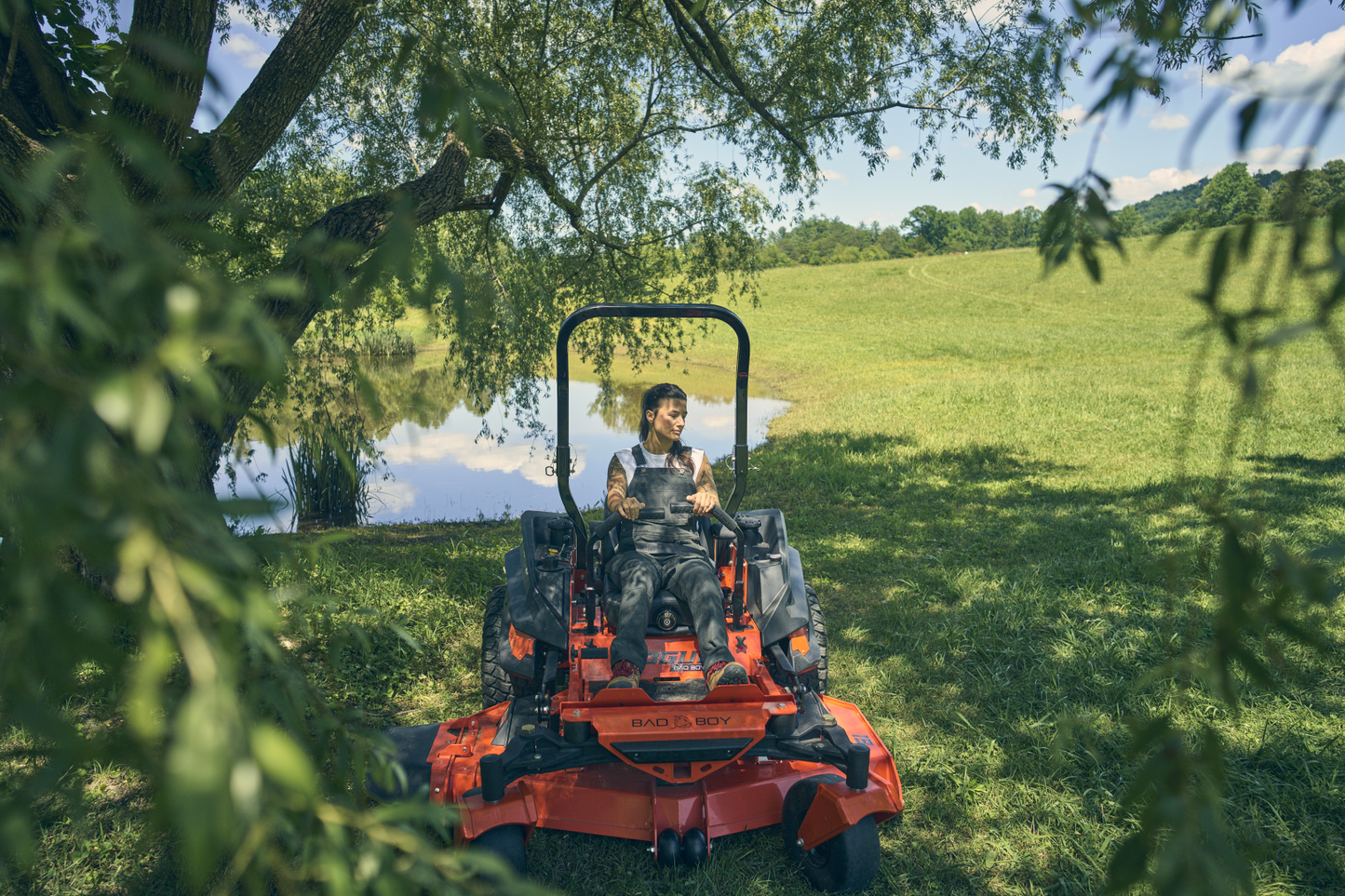 Person operating a red lawn mower in a grassy field with trees and a pond in the background
