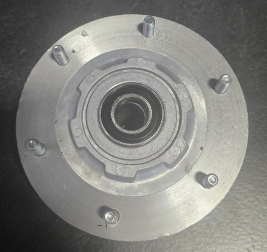 A circular metallic silver bearing housing with a central bearing and four visible mounting bolts, resting on a dark textured surface.