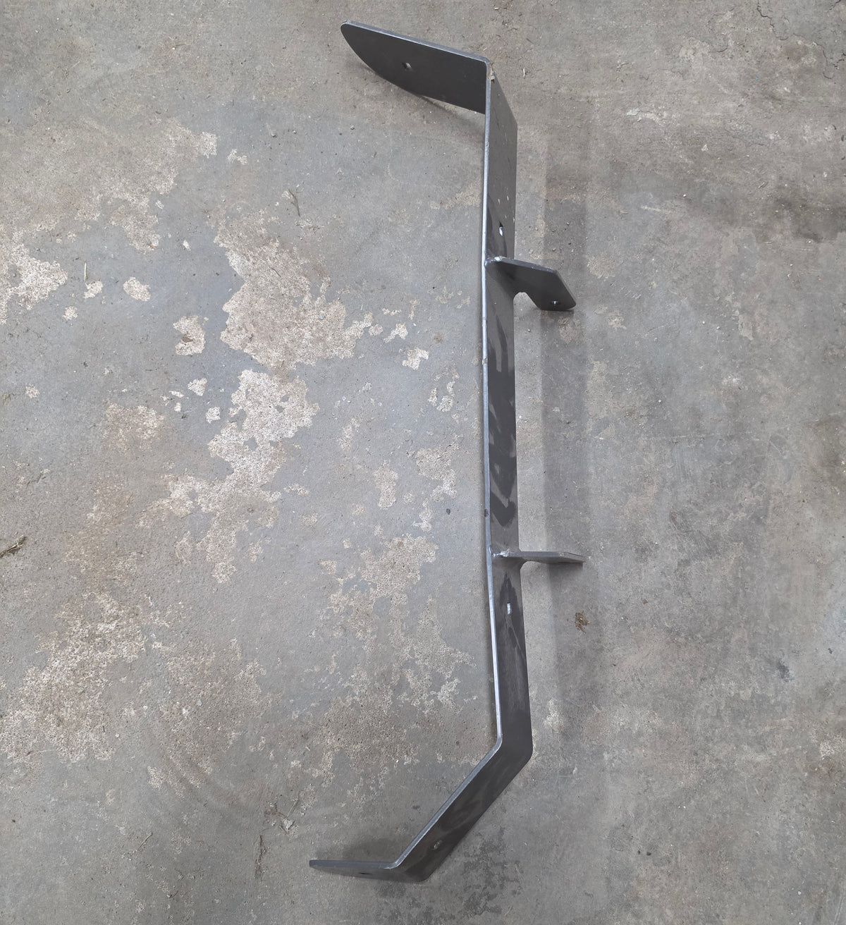 A dark gray metal bracket or support arm with multiple welded joints, photographed on a worn concrete floor.