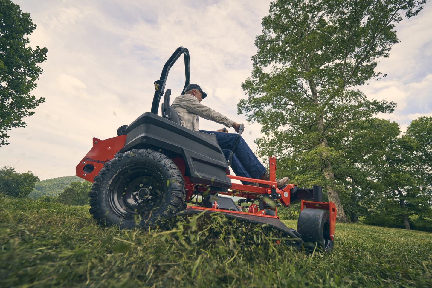 Person operating a red lawn mower in a grassy area with trees and a cloudy sky.
