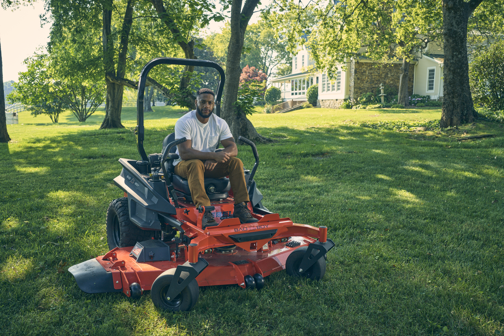 Person sitting on a lawn mower in a grassy yard with trees and a house in the background