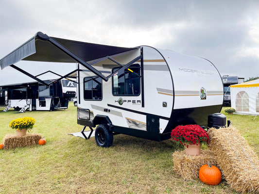 Small travel trailer with an extended awning at a campground, decorated with pumpkins and flowers.