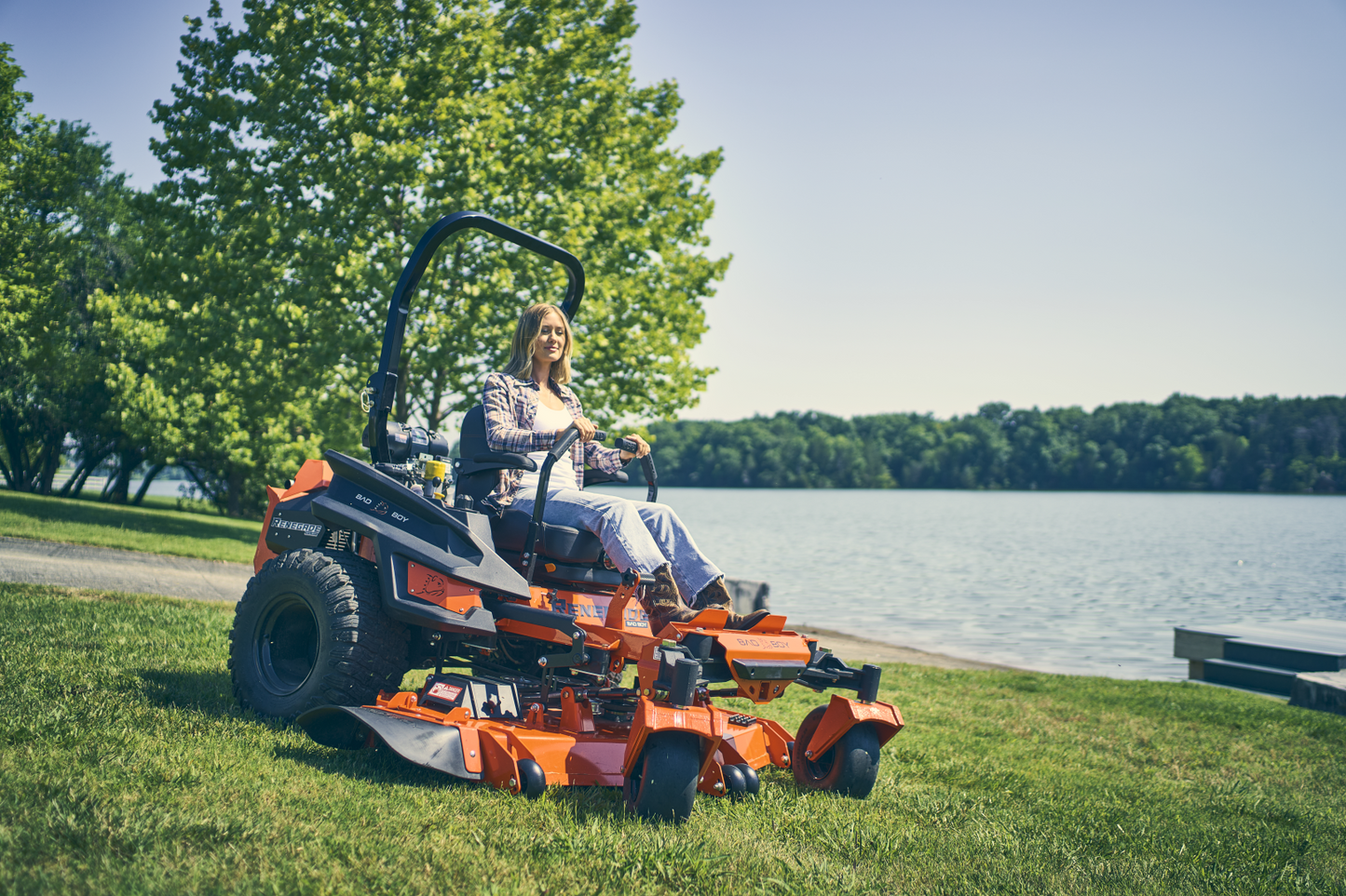 Person operating a lawn mower by a lake with trees in the background