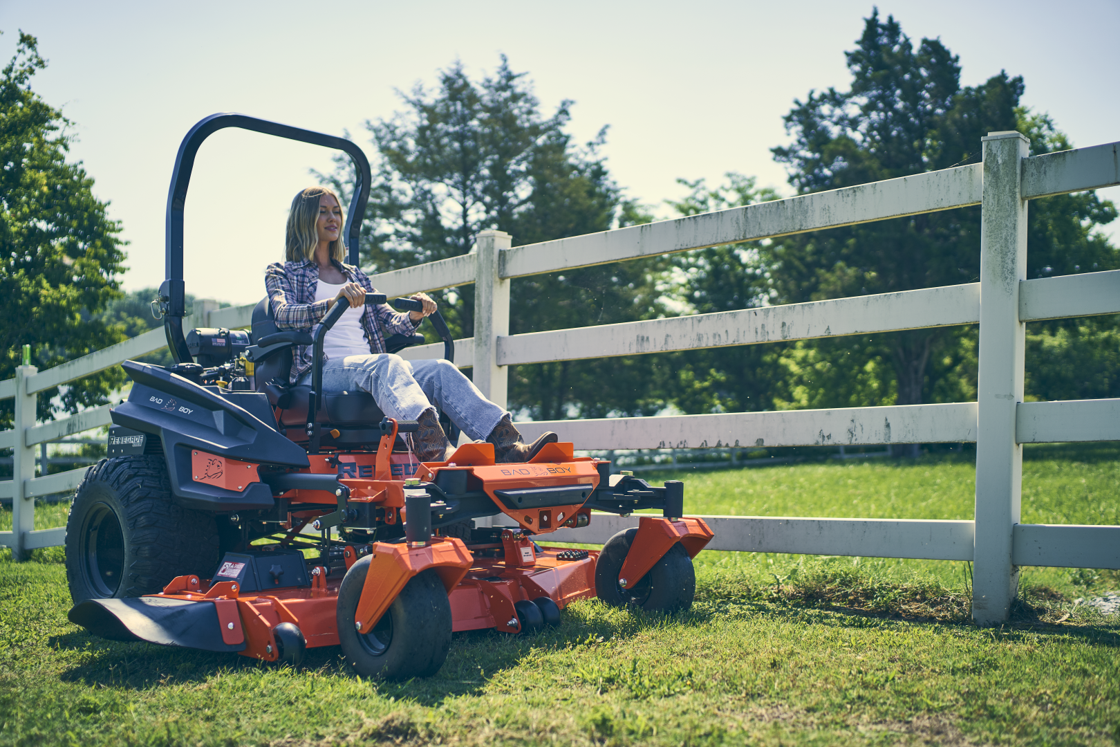 Person operating a large orange lawn mower in a grassy area with a white fence and trees in the background.