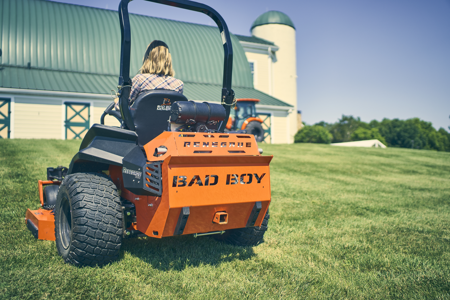 Person operating an orange Bad Boy mower on a grassy field with a barn and silo in the background.