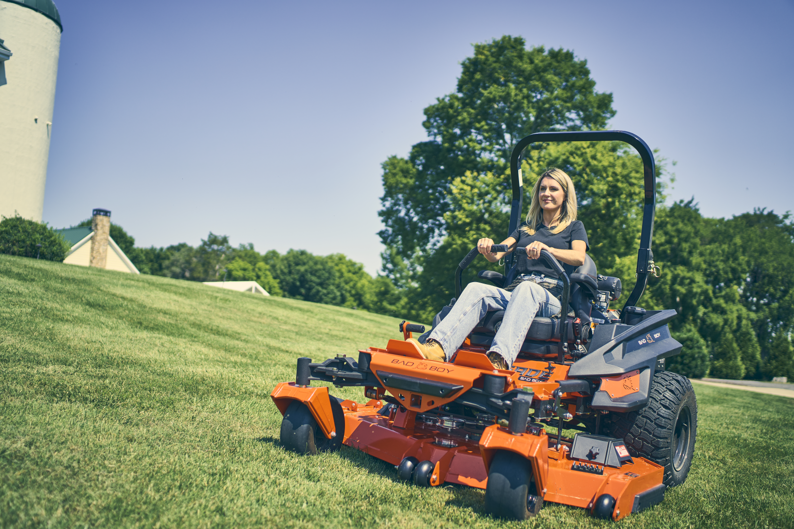 Person operating a large orange lawn mower on a grassy area with trees and a building in the background.