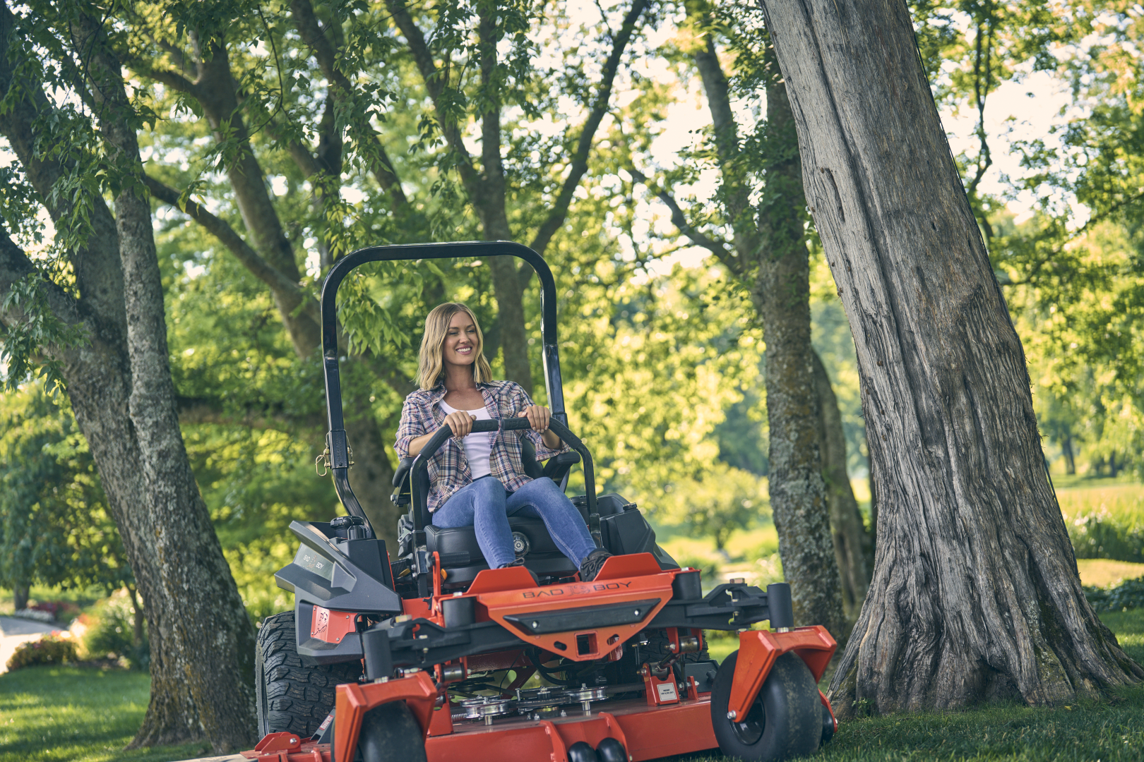 Woman operating a red riding lawn mower in a park with trees and greenery.