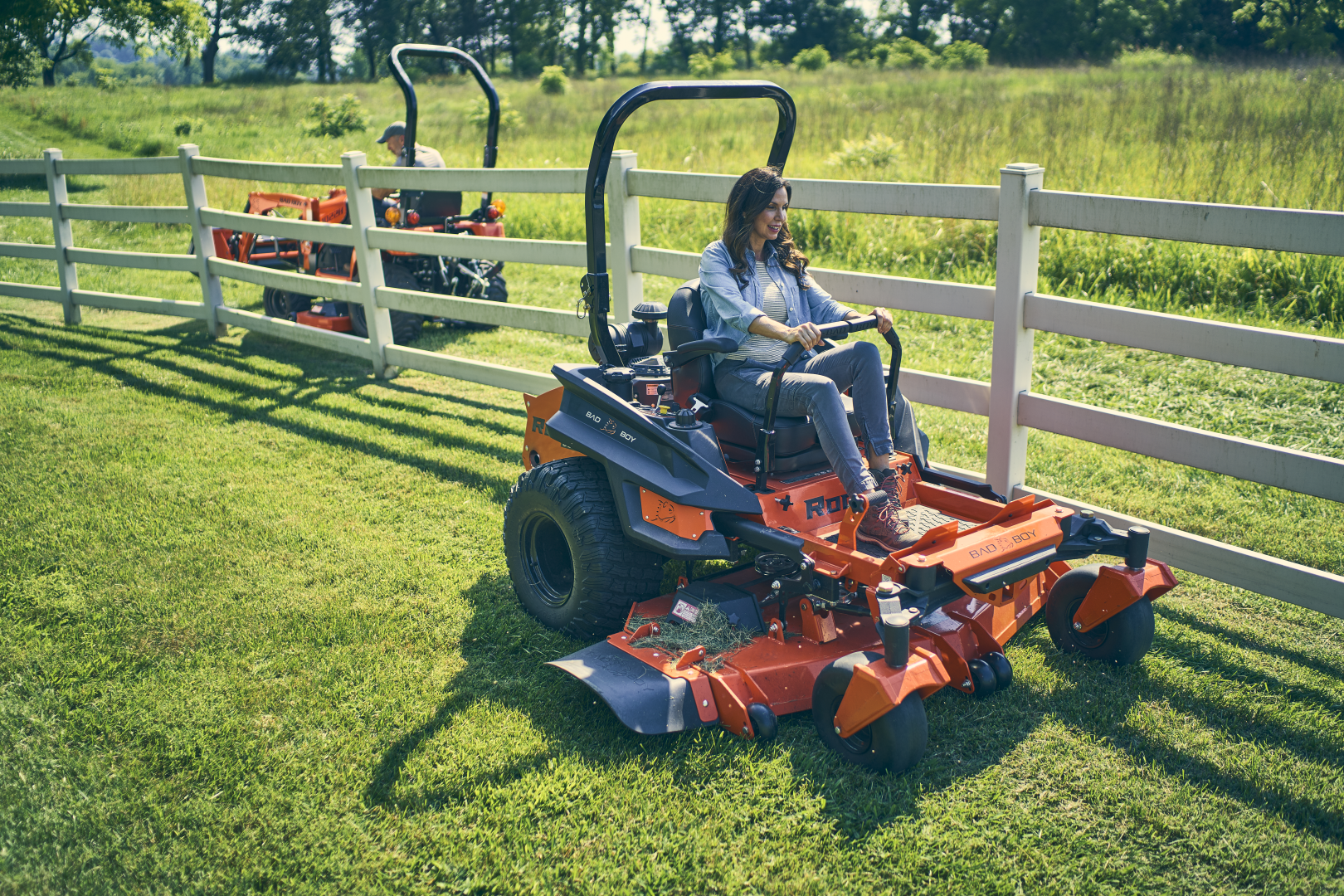 Two people operating a large orange lawn mower in a grassy field with a white fence.