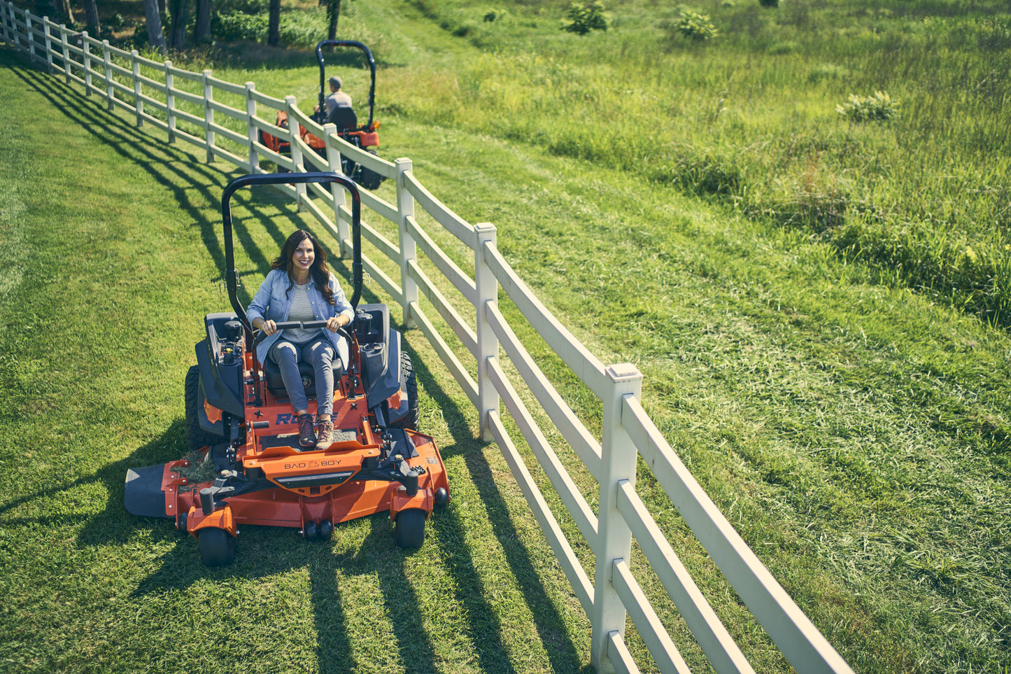 Two people using a lawn mower on a grassy field with a white fence in the background.