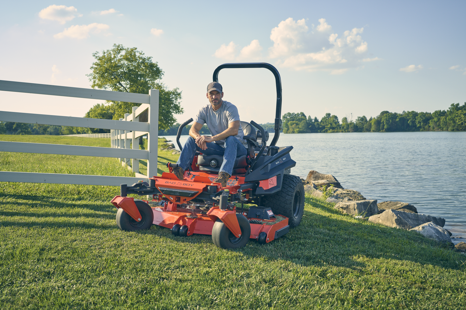 Man sitting on a red riding lawn mower by a lake with a white fence in the background.