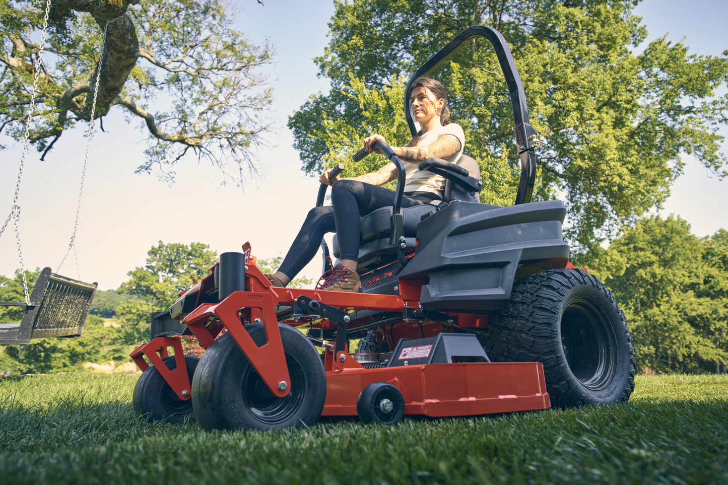 Person operating a red zero-turn lawn mower in a grassy area with trees in the background