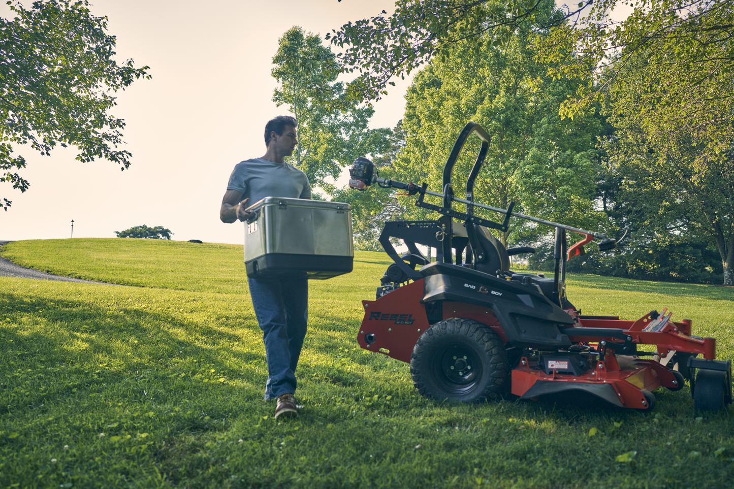 Person carrying a large metal box next to a lawn mower on a grassy area with trees in the background