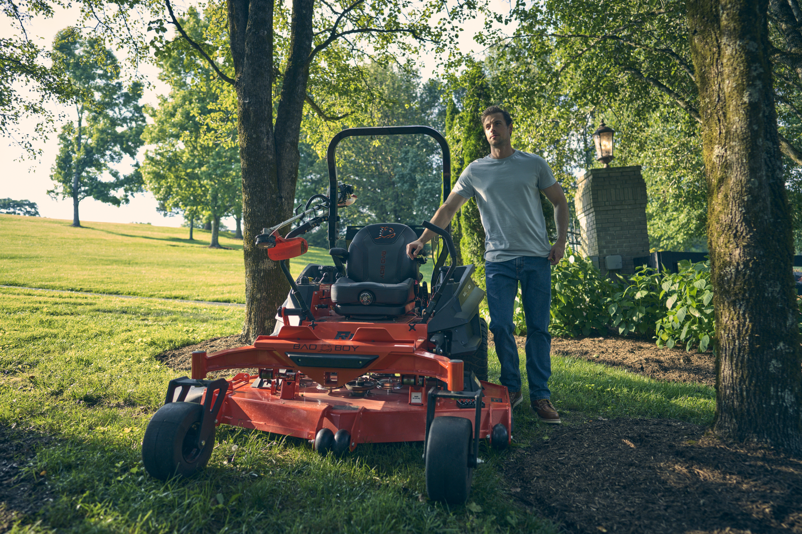 Man standing next to a red lawn mower in a park-like setting with trees and grass.