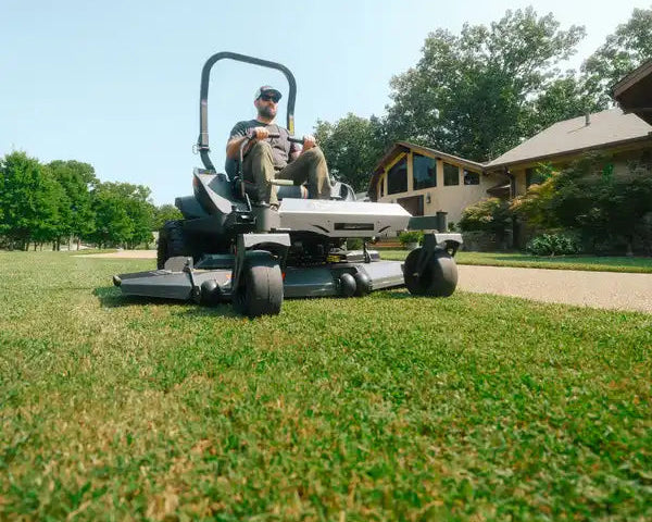 Person operating a riding lawn mower on a grassy area with trees and a house in the background.