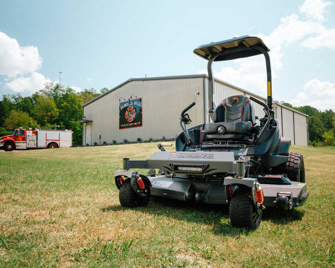 Large lawn mower on a grassy field with a building and truck in the background