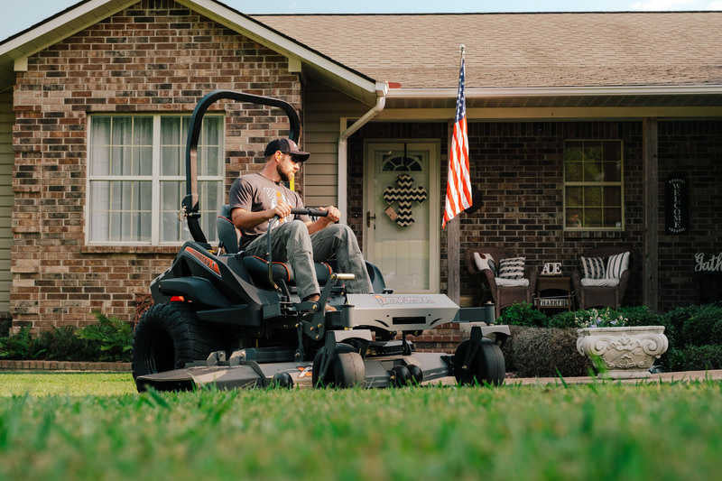 Man operating a riding lawn mower in front of a house with an American flag.