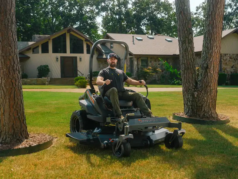 Man operating a riding lawn mower on a grassy area with trees and a house in the background.