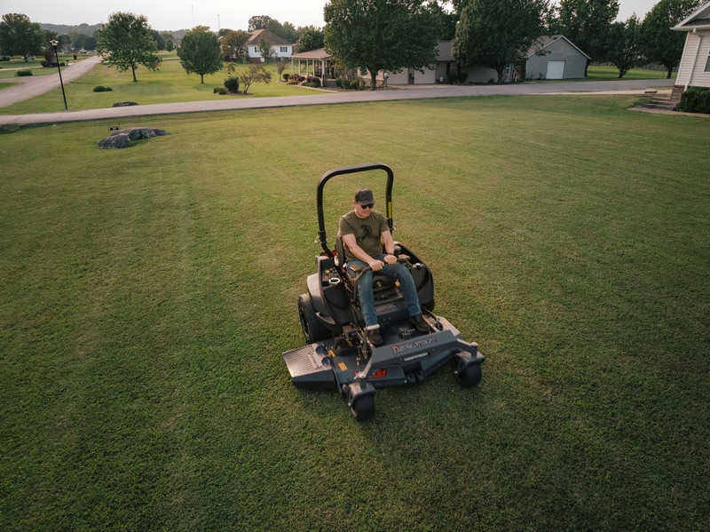 Person operating a riding lawn mower on a green lawn with houses and trees in the background.