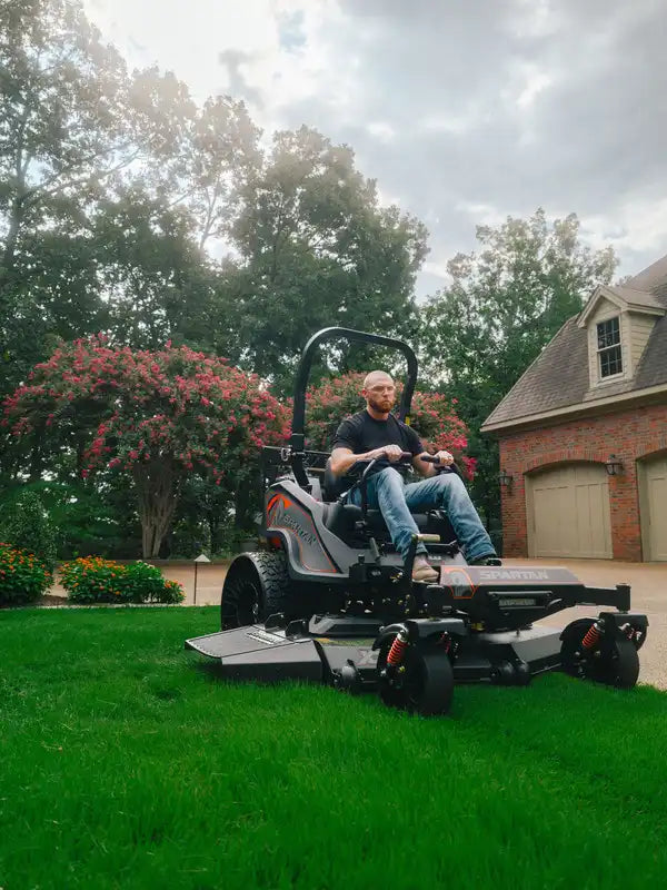 Person operating a large lawn mower on a green lawn with trees and a house in the background.