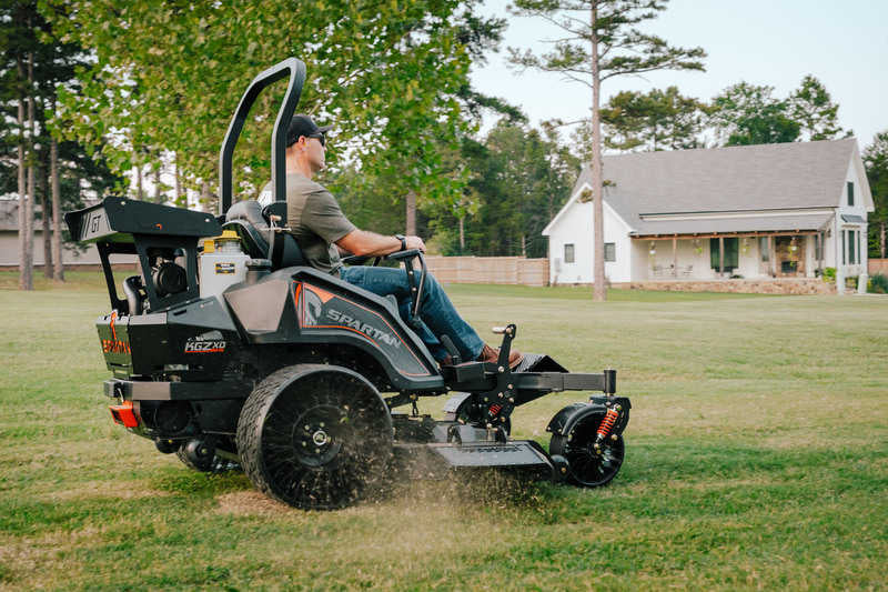 Person operating a zero-turn lawn mower in a grassy area with trees and a house in the background.