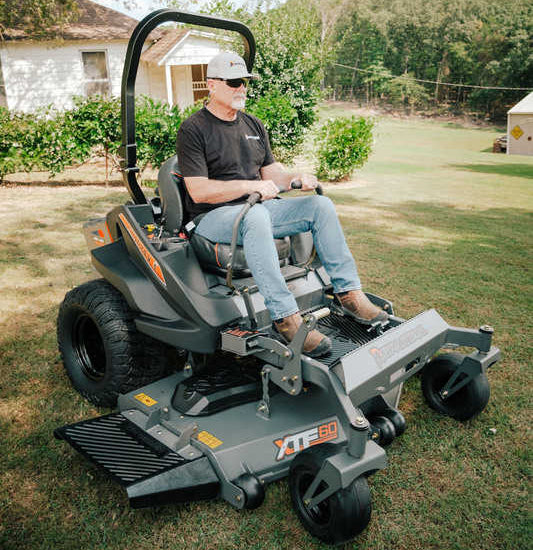 Person operating a large lawn mower on a grassy area with trees and a building in the background.