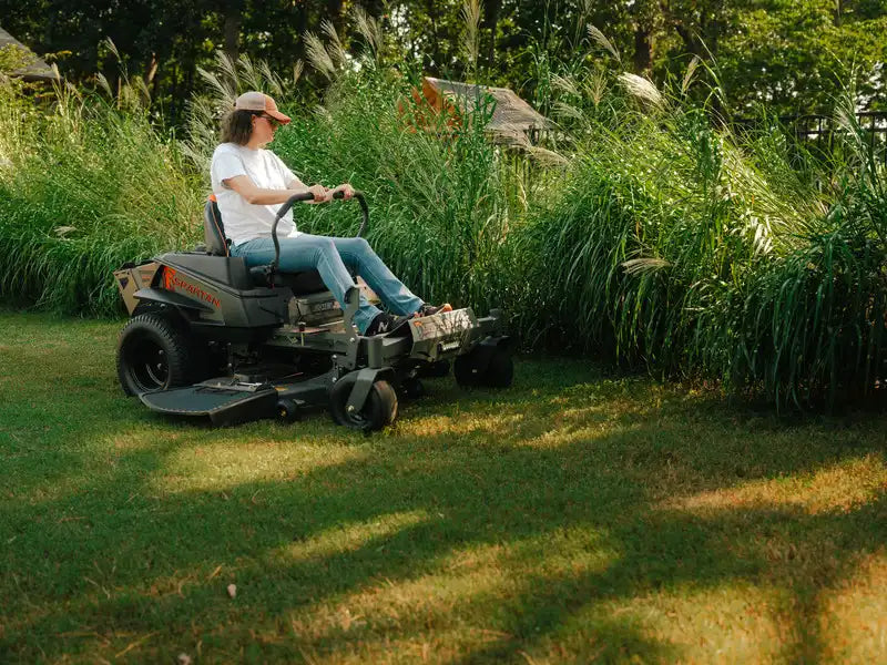 Person using a riding lawn mower in a grassy area with tall plants.