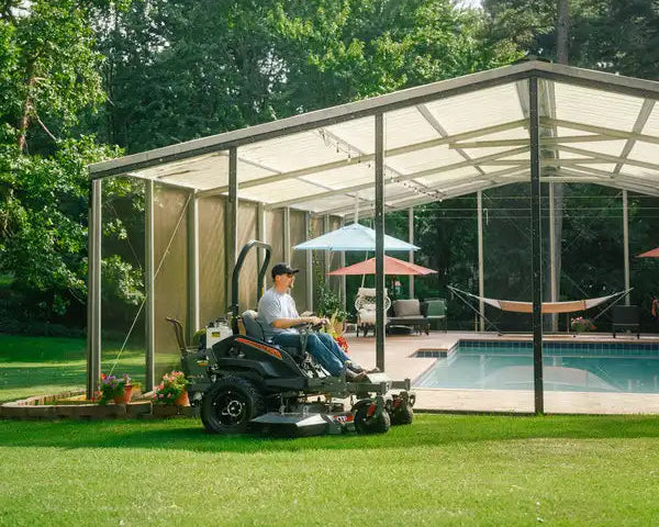Man sitting on a lawn mower in a modern glass house with a pool and garden.