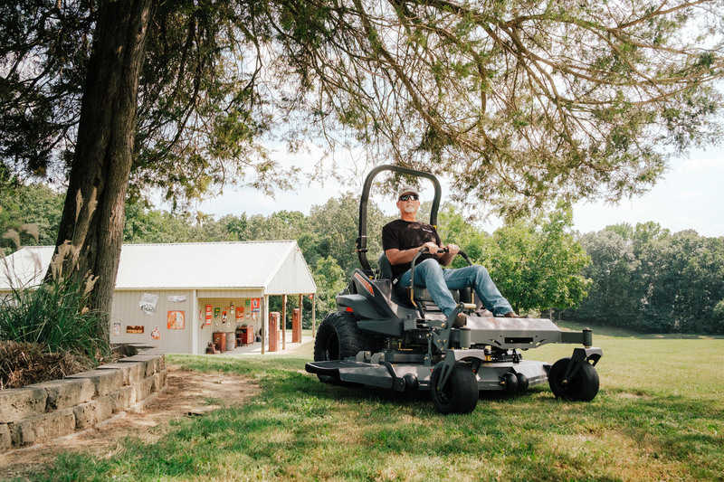 Man operating a zero-turn lawn mower in a grassy area with trees and a building in the background.