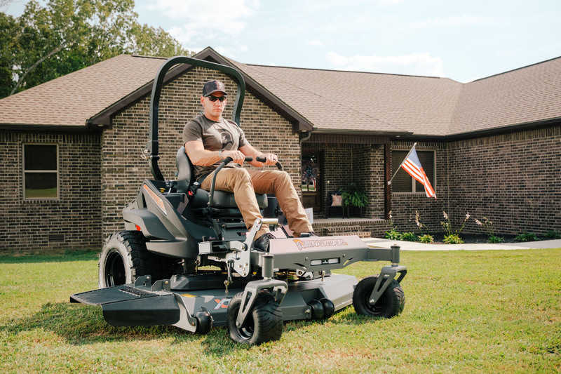 Man operating a large lawn mower in front of a house with an American flag.