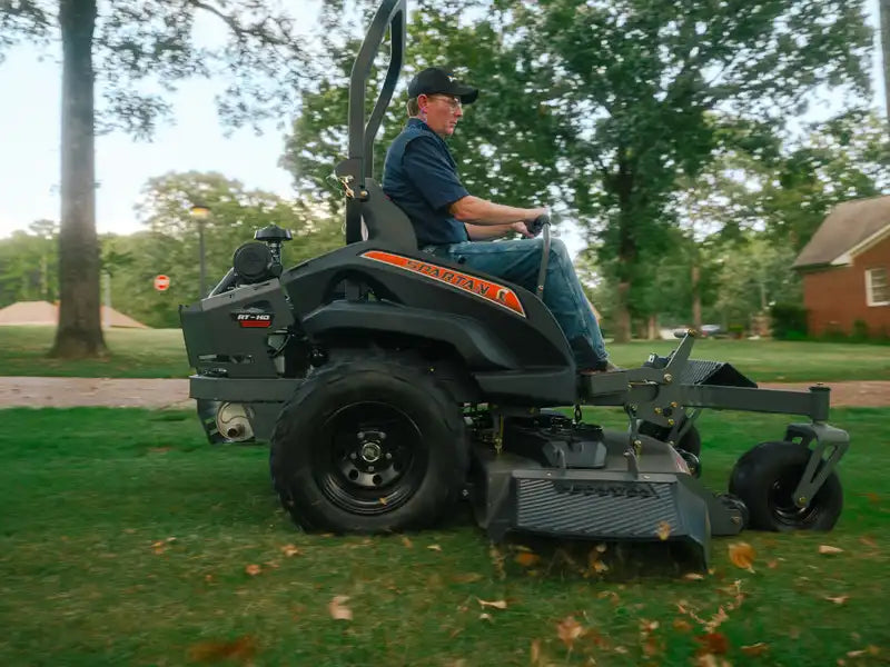 Man operating a riding lawn mower in a grassy area with trees and a house in the background.