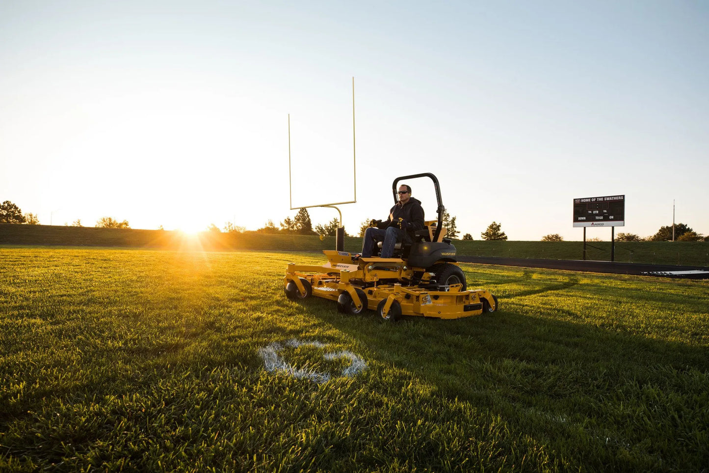 Person operating a lawn mower on a sports field with sunset in the background