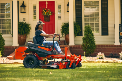 Person using a riding lawn mower in front of a house with a red door.