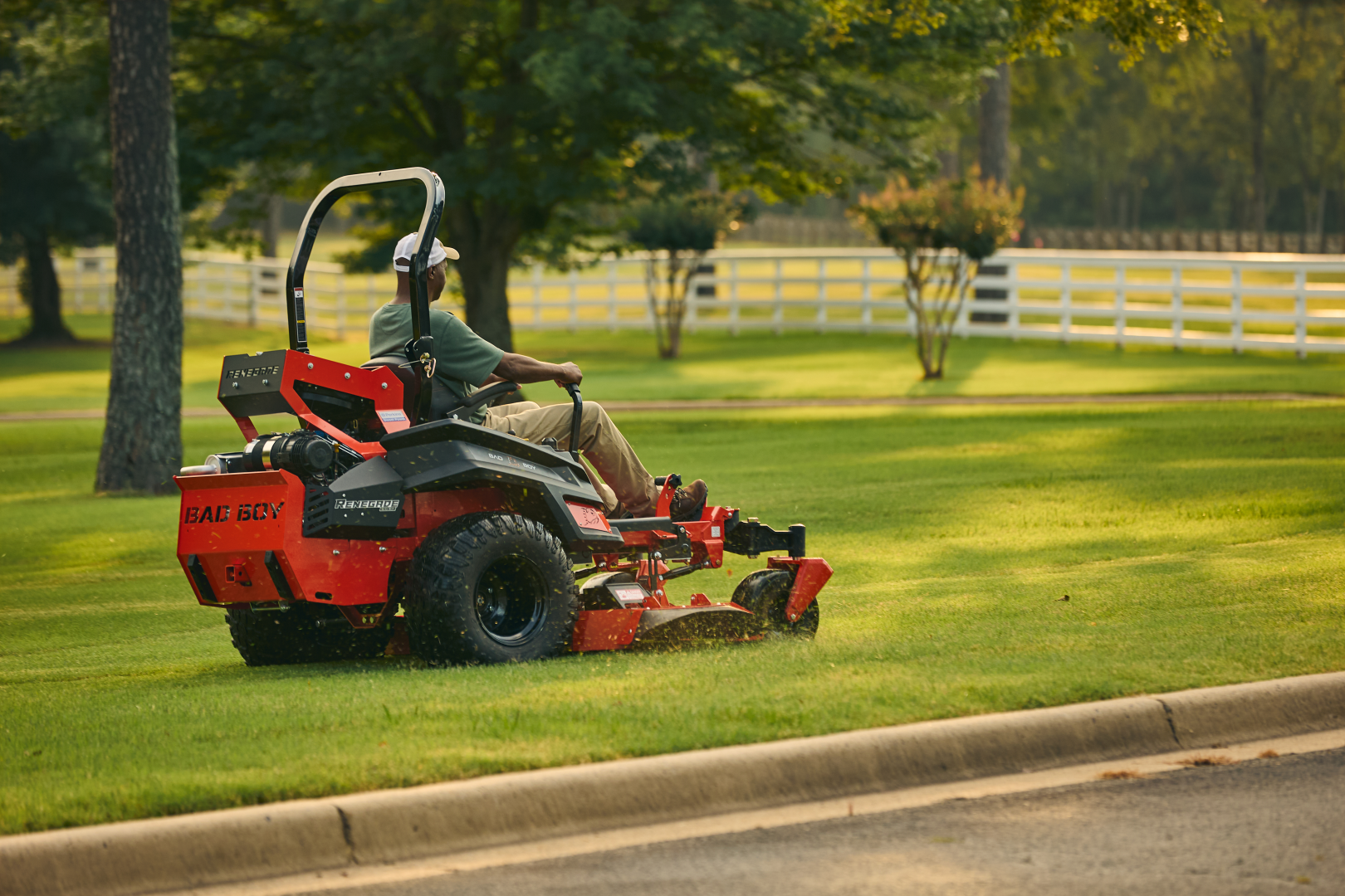 Person operating a red riding lawn mower on a green lawn with trees and a white fence in the background.