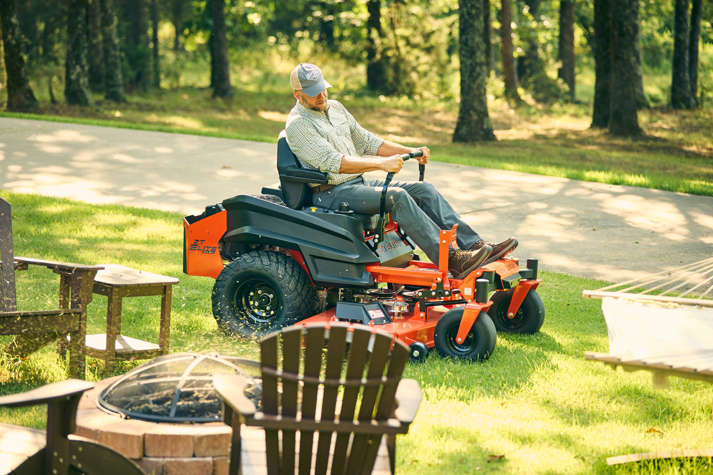Man operating a riding lawn mower in a backyard with trees and a fire pit.