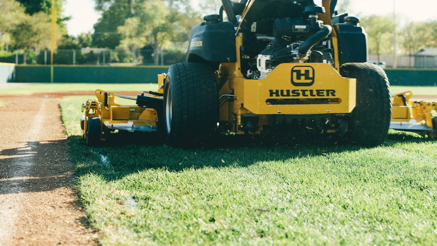 Yellow Hustler zero-turn lawn mower cutting grass on a dirt path.