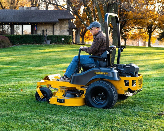 Man operating a yellow HUSTLER lawn mower on a grassy lawn with trees and a building in the background.