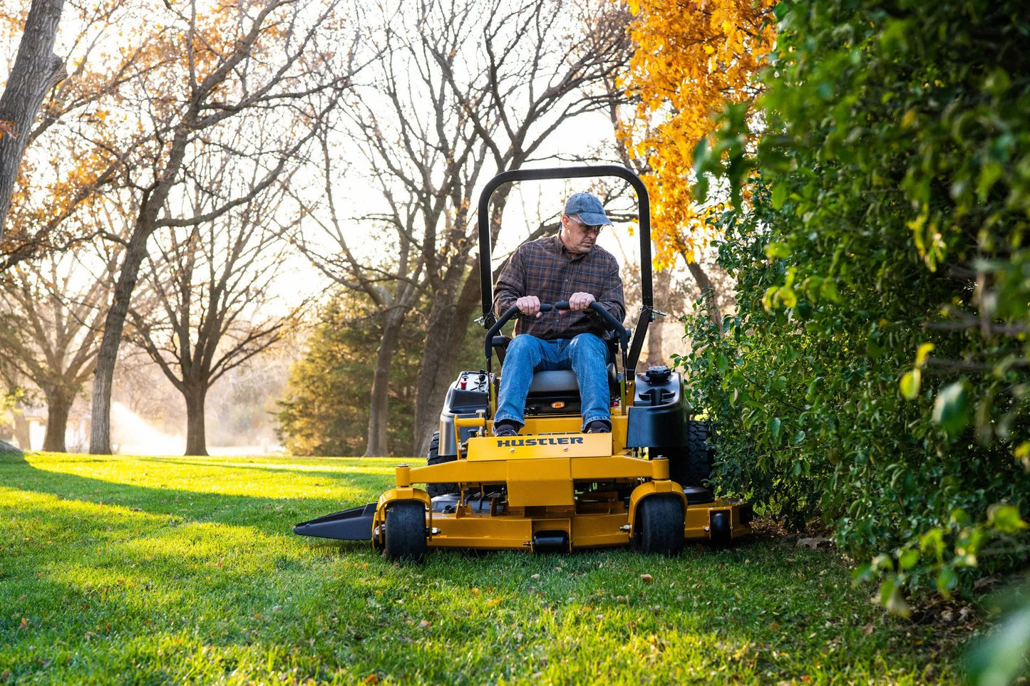 Person operating a yellow Hustler mower in a park setting with trees and grass.