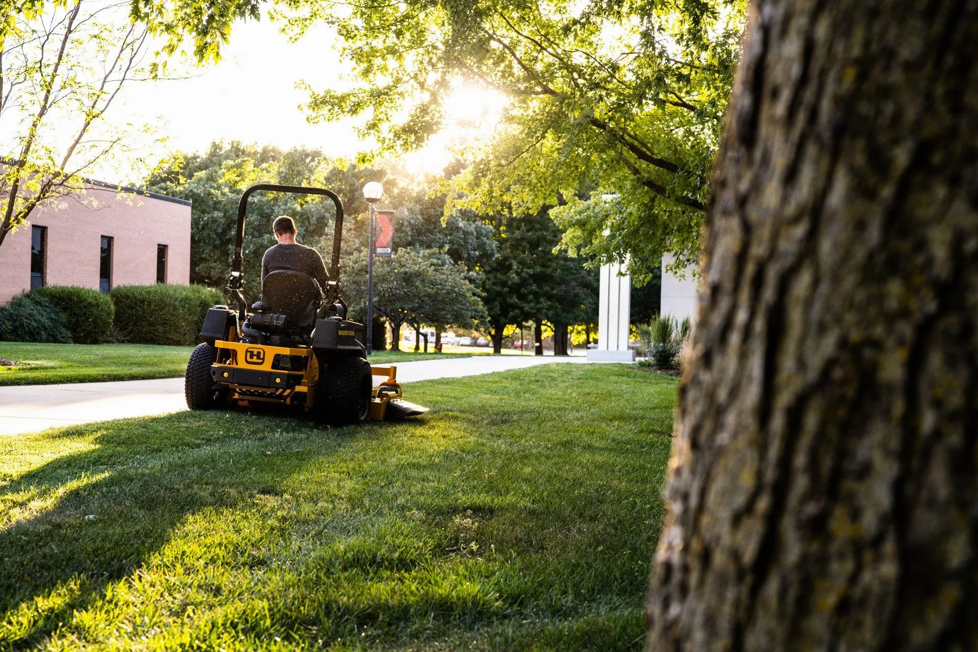 Person operating a lawn mower in a park-like setting with trees and buildings in the background.