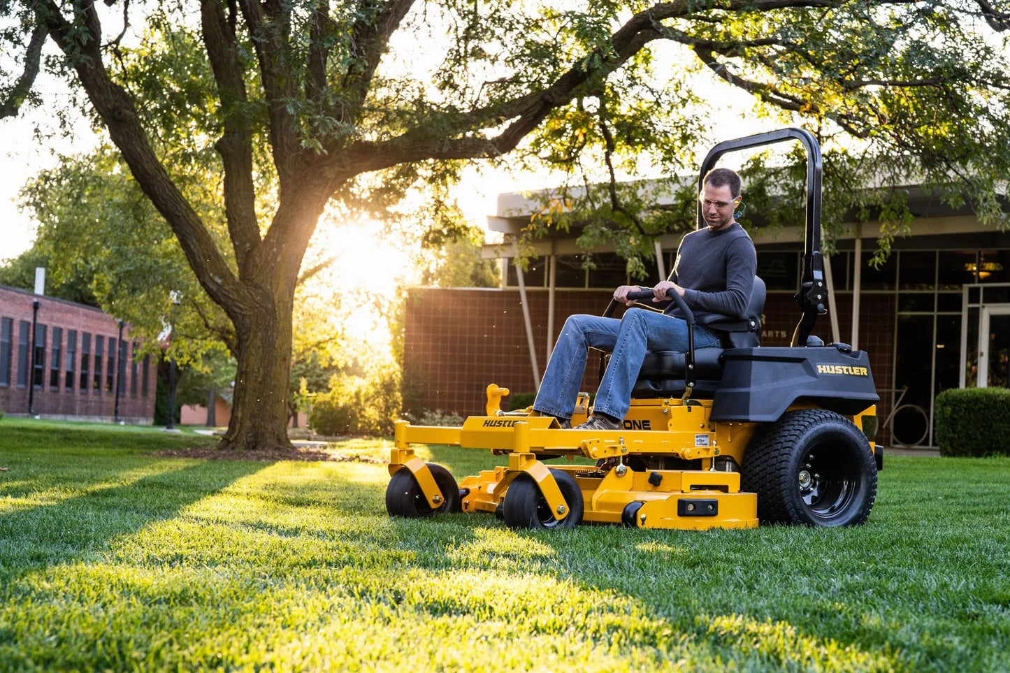 Man operating a yellow riding lawn mower in a park setting with trees and buildings in the background.