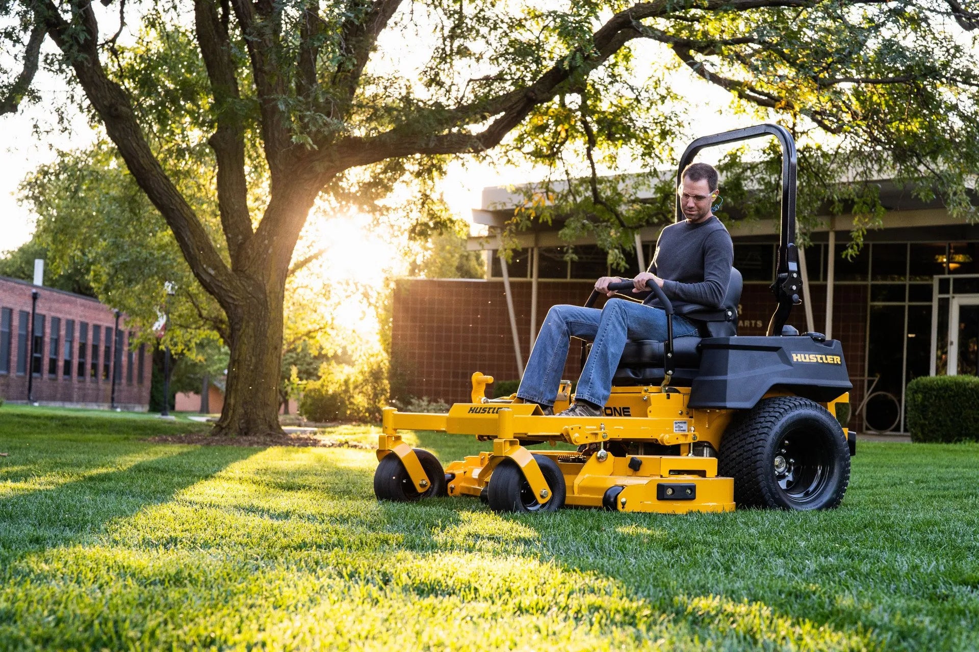 Man operating a yellow riding lawn mower in a park setting with trees and buildings in the background.