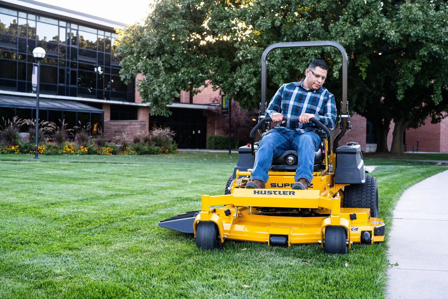 Man operating a yellow Super Mower on a grassy area with trees and a building in the background.