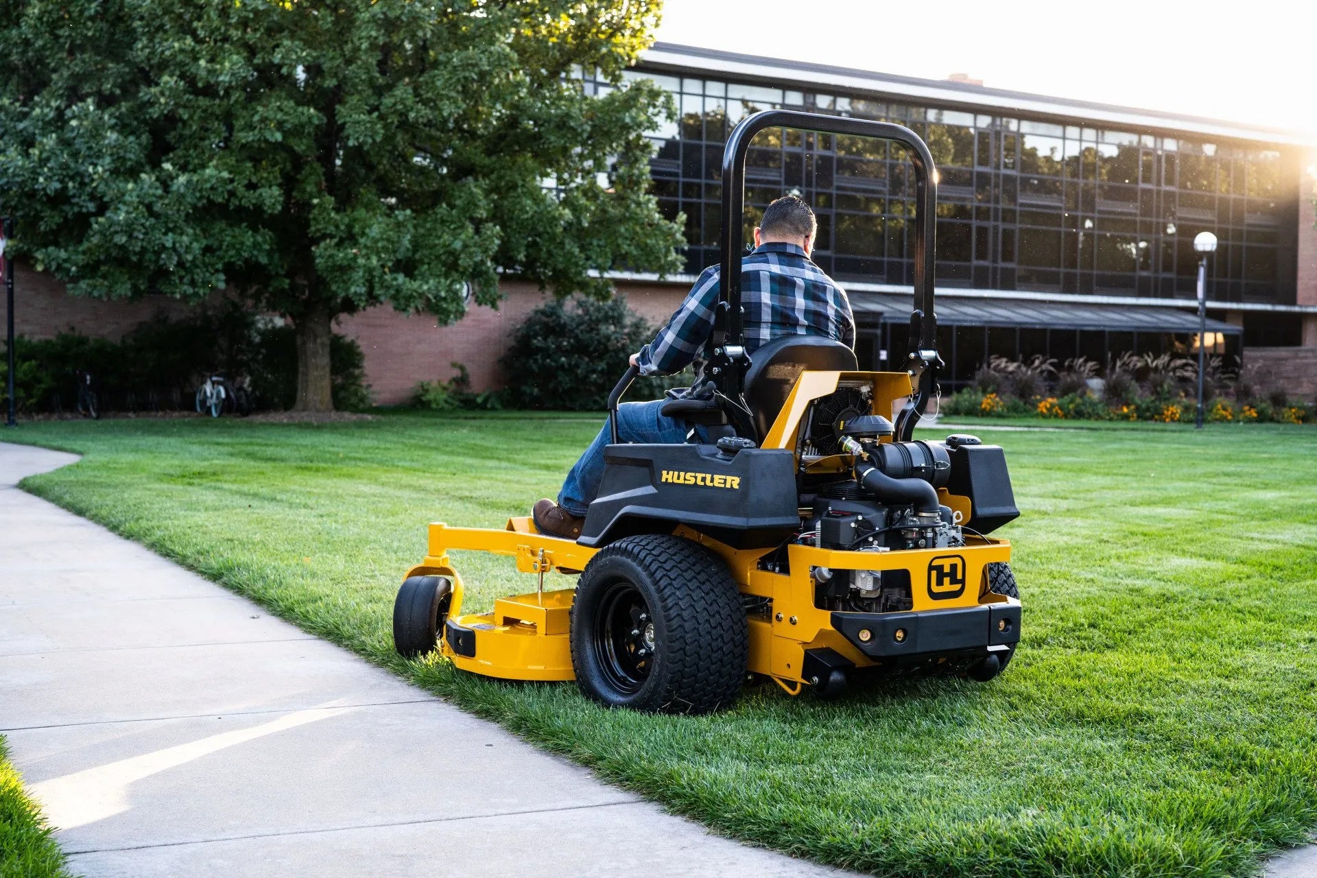 Person operating a Hustler zero-turn lawn mower on a grassy area with a building in the background.