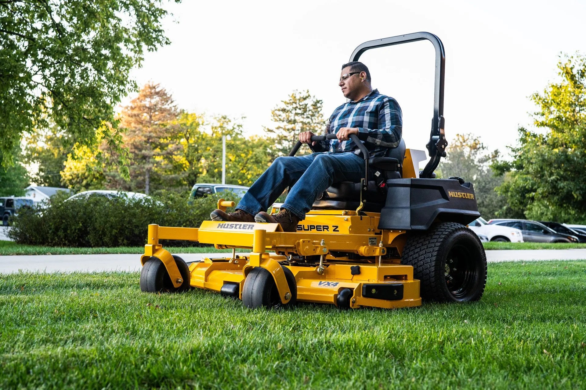 Man operating a yellow riding lawn mower in a park-like setting with trees and cars in the background.