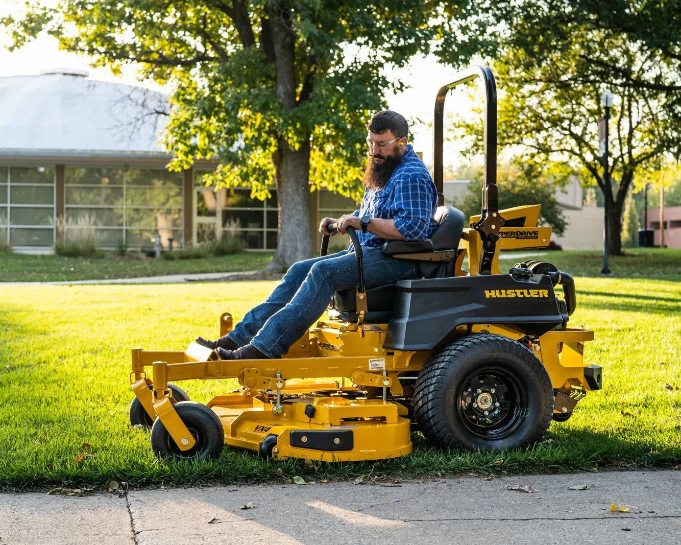 Man operating a yellow riding lawn mower on a grassy area with trees and a building in the background.