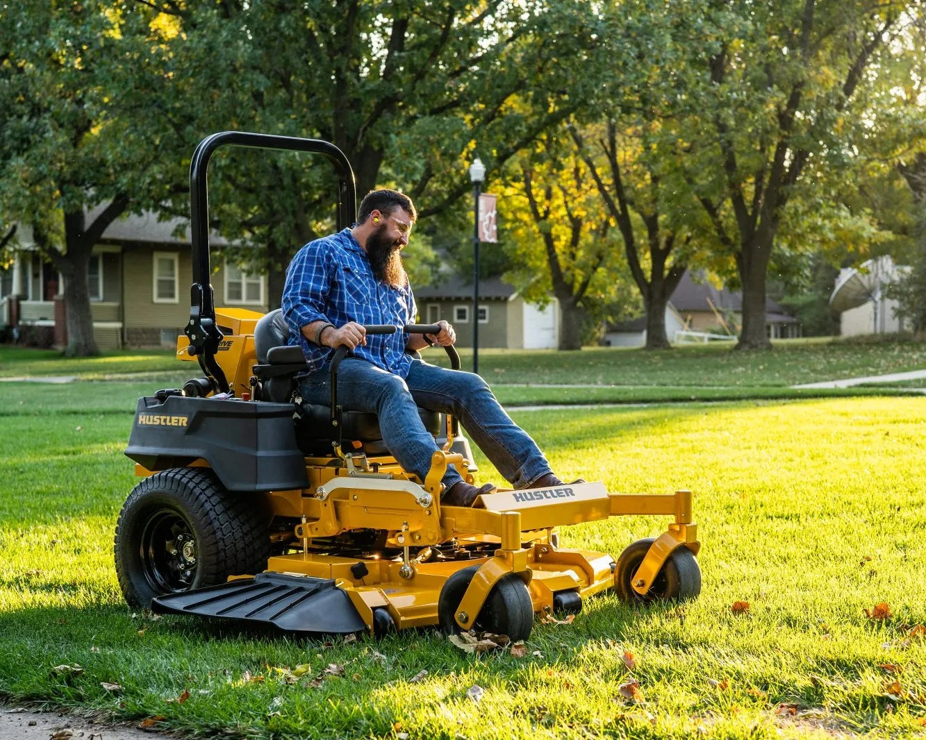 Person operating a yellow riding lawn mower on a grassy area with trees and houses in the background.