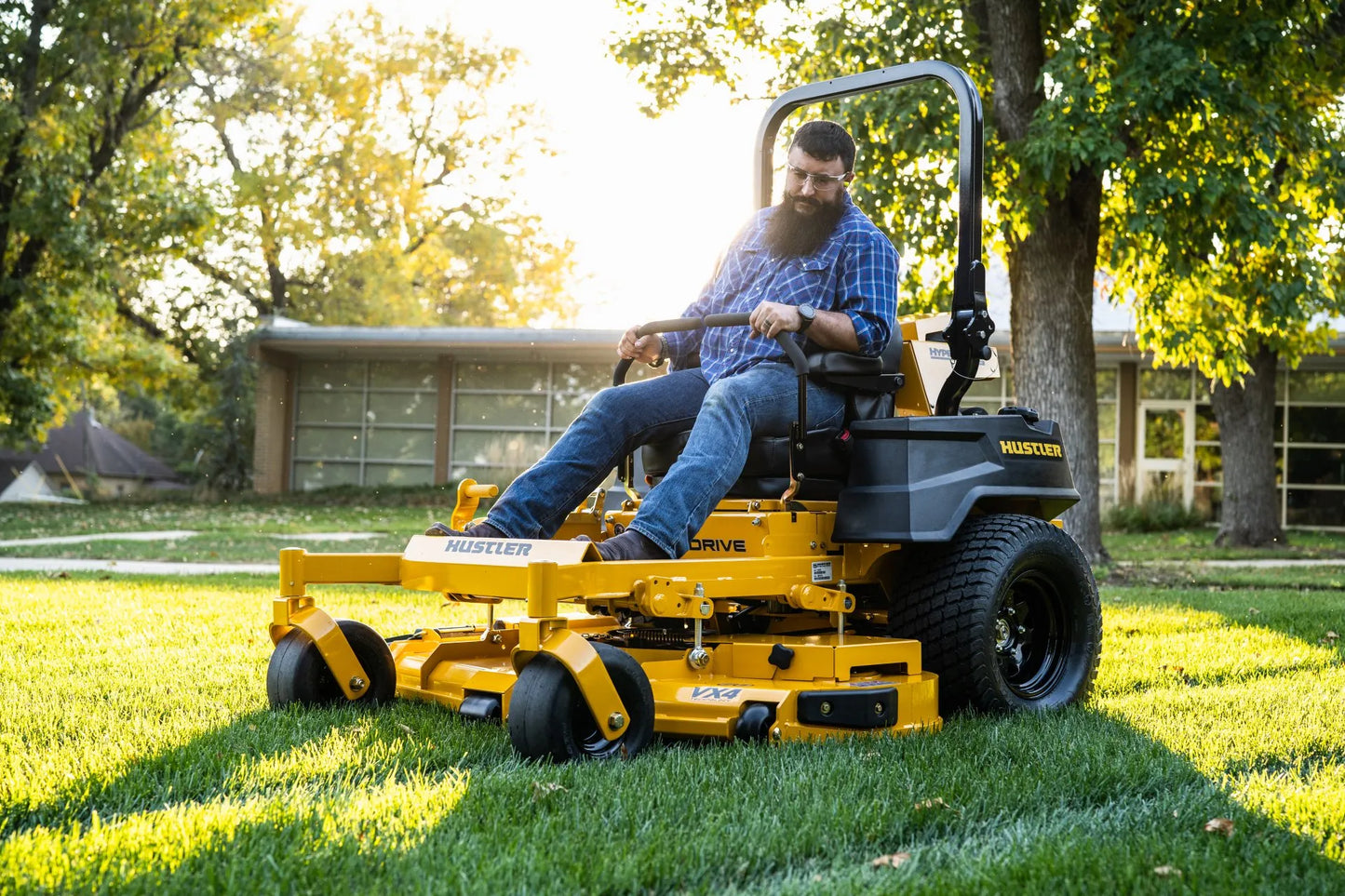 Man operating a yellow riding lawn mower in a grassy area with trees and a building in the background.